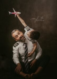 Bambino in braccio al papà pilota solleva un aeroplanino verso l'alto mentre il papà lo guarda con una smorfia divertita